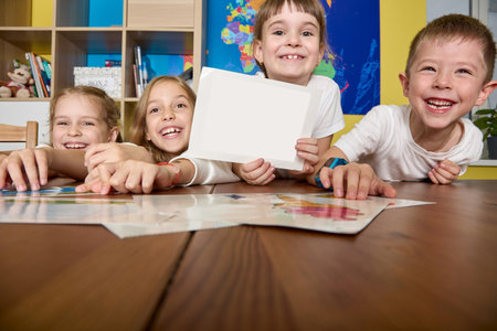Happy children in the background of world map sit at the table, study with cards. Fun educational activity for young children in an educational institution. Girl demonstrates the place for layoutの写真素材