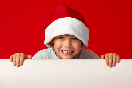 Joyful handsome boy smiling holds his hands on white billboard. Close up of boy in red New Years hat smiling looking at the camera next to billboardの写真素材