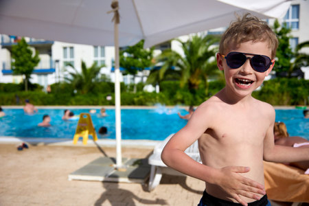 Cheerful boy in sunglasses basks in the sun next to a protective umbrella after swimming in the pool. Smiling child after swimming dries under the warm bright sunの写真素材