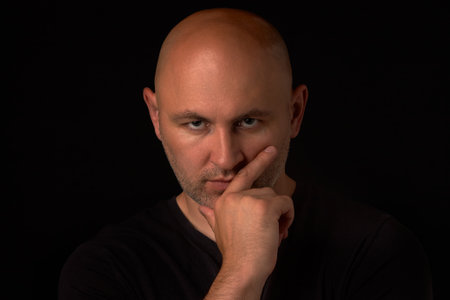 Bald, bearded man looks thoughtfully into the camera and holds his hand on his chin, on a black background. A man in depression, a midlife crisisの写真素材