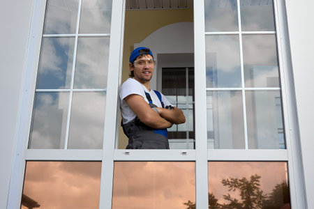 Portrait of a young worker in a work uniform and a blue cap standing with smile at the window,...