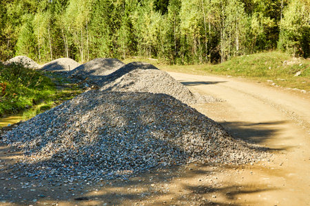 Large piles of rubble are laid out on the side of a country road through the forest to repair the road. Dirt winding road through the forest with scattered piles of gravelの写真素材
