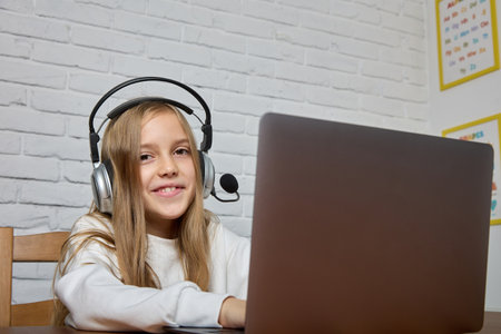 Teenage girl with long hair sits with a headset on her head at a table and a laptop in a classroom,...