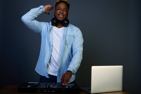 Black man DJ in a blue shirt makes music on his vibe. In the studio with headphones, a mixer and other equipment. Enjoys his occupation. An active African-American with vivid emotions.の写真素材