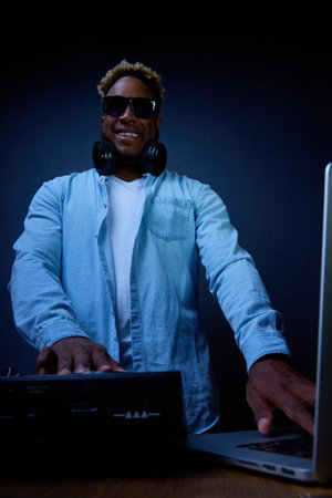Smiling fashionable dark-skinned guy in sunglasses posing at the DJ console. young African American man in DJ headphones at a mixing console on a gray background is spinning tracksの写真素材