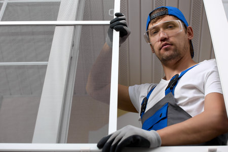 View from below of a professional installer in a work uniform, standing by the window in protective glasses and gloves. Master installer poses at the window holding a masked insect net with his handの写真素材