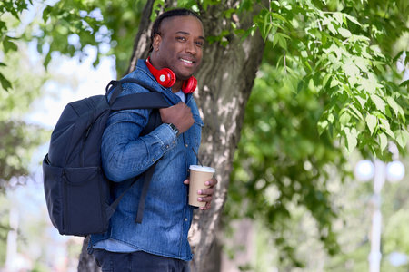 Dark-skinned man with heavy backpack behind his back, smiling, stands in shade of trees in green...