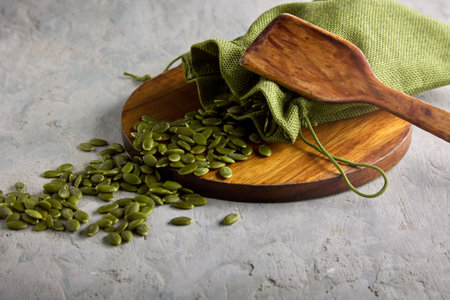 Roasted pumpkin seeds on a wooden stand along with a bag of wooden spoon on a gray background. Raw green pumpkin seeds on a round wooden plate are laid out chaoticallyの写真素材