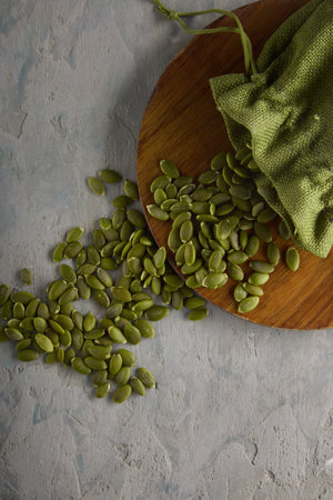 Raw peeled pumpkin seeds on a wooden board fell out of a green bag, top view. A scattering of useful pumpkin seed kernels on a gray background with a storage bagの写真素材
