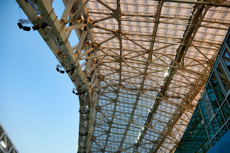 Bottom view of the stadium roof with cloudless blue sky. Floodlights are placed high on the metal...