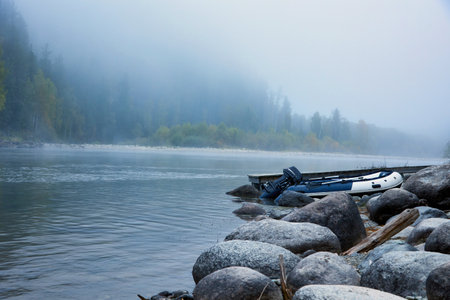 Morning autumn landscape of mountain river or lake, fog descends from the mountains. On the bank of a mountain river with a creeping fog, large stones and an inflatable boat with an outboard motorの写真素材