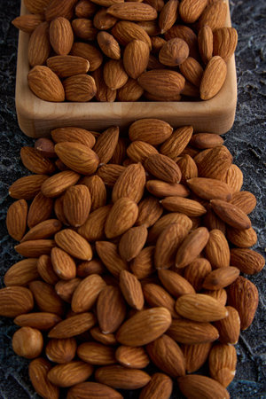 Delicious roasted almonds close-up on a gray textured background are scattered from a square-shaped wooden plate. Raw peeled almonds stacked in a heap on the table, top viewの写真素材