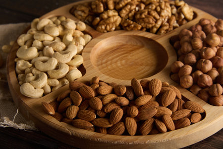 Wooden round plate with various types of nuts, standing on a burlap on the table. Dried cashews, almonds, hazelnuts and walnuts are laid out on a wooden rackの写真素材