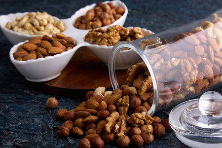 On a gray table, nuts are laid out on a beautiful plate next to a scattered mixture from a glass jar. Assorted nuts in a dried fruit store on a gray textured backgroundの写真素材