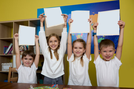 Satisfied children stand against background of yellow wall in office raised their hands up with white sheets of paper. Schoolchildren stand with paper in their hands raised at desk in the classroomの写真素材