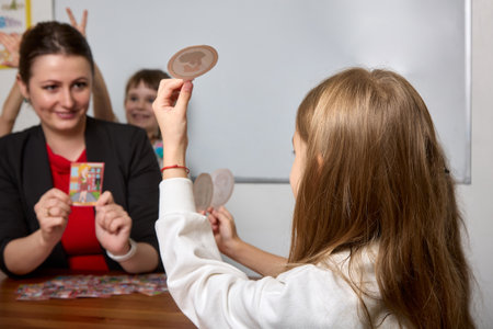 In the classroom, against the background of a white board, the teacher sits at the table showing the children educational cards. Educational activity of children in a modern schoolの写真素材