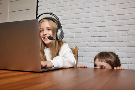 Little girl watches her older sisters online class. In a room with a white brick wall, a teenager sits at a table in a headset with a laptop, next to it is a small child watchingの写真素材