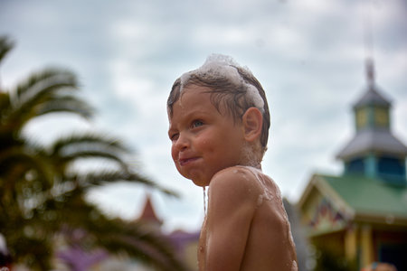 Funny little boy with foam on his head puffed up his cheeks, closed one eye, had fun at a foam party. Happy cute kid at a foam party on the beachの写真素材