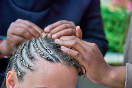 An African master with an assistant in 4 hands carefully and quickly braid a young woman Afrocos. Process of weaving a popular youth hairstyle by two experienced hairdressersの写真素材