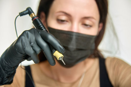On blurry background, face of a permanent makeup artist in a black protective mask performing work, holding the device in his hand. Close-up of tattoo tool in hand of an experienced cosmetologistの写真素材