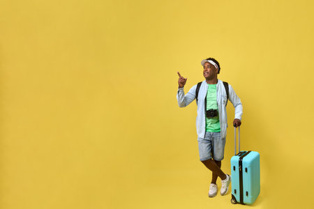 Happy young man standing on a yellow background with a suitcase pointing to an empty space for a travel agency advertisement. A cheerful traveler with luggage is going on summer vacationの写真素材