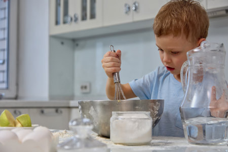 Focused boy carefully mixes the ingredients for the cake in a metal dish in the home kitchen. Young independent child cook learns to knead the dough with a metal whiskの写真素材