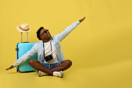Young African man in sunglasses and denim shirt sits next to suitcase with arms outstretched to sides like wings of airplane. Positive traveler dreams, took pose of a flying plane before departureの写真素材