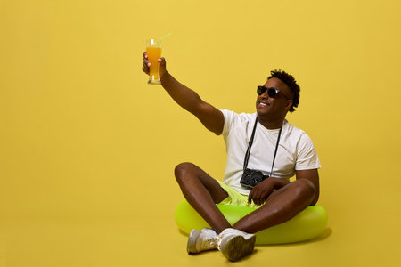 Delicious refreshing cocktail in a glass glass was lifted up by an African young man sitting in a swimming circle. African-American tourist relaxing on the beach, drinking a healthy drink on vacationの写真素材