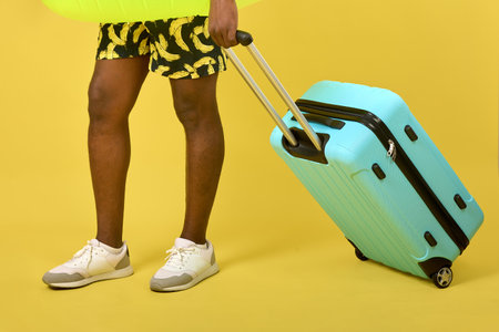 An African man in shorts with a banana pattern on a yellow background rolls a large blue suitcase on...