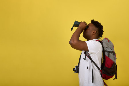 An adult African guy in a white T-shirt with a backpack watching through binoculars. African-American male tourist traveling with a backpack, exploring the area with binocularsの写真素材