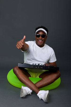 Modern African-American DJ smiles, pointing his finger up, playing music on a gray background. Joyful black man in white T-shirt and sunglasses is playing music while sitting in an inflatable circle.の写真素材