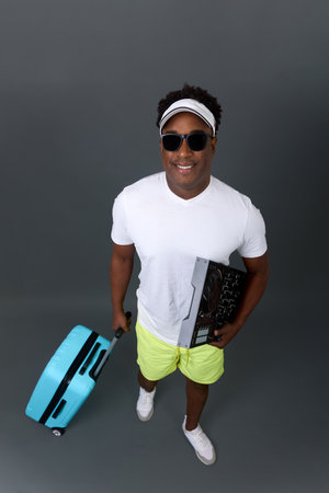 Handsome young black man tourist in sunglasses with a suitcase and equipment on a gray background...