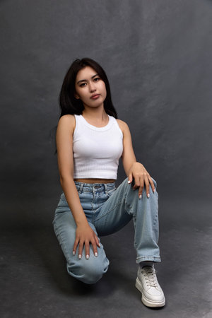 Serious and calm young woman of Asian nationality sat down on one knee in fashionable jeans. Stylish young girl in modern clothes confidently holding on to the cameraの写真素材
