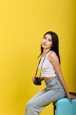 Young Asian brunette on a yellow background sitting on a suitcase with a camera hanging around her neck. Long-haired brunette in jeans with a kind look look at the cameraの写真素材