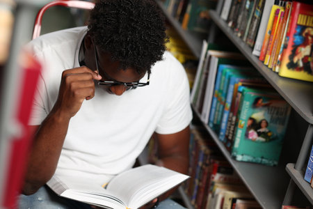 Young african male student enthusiastically reads new book while sitting stepladder among shelves in library. Young curly-haired dark-skinned guy in white t-shirt reading interesting book in bookstoreの写真素材
