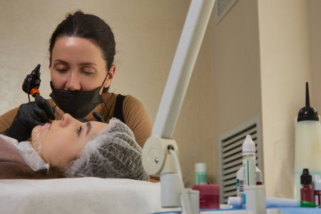 A professional master of a cosmetology clinic performs the procedure of permanent makeup of a young...