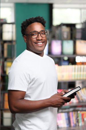 Young black man smiling contentedly and looking at camera while standing among store bookshelves....