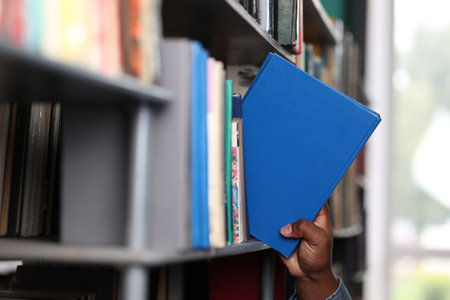 Close-up of a black mans hand taking out a blue-bound book from a bookshelf in a library. Educational literature is used in an international university by an African studentの写真素材
