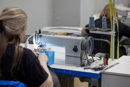 A seamstress in casual clothes sits at a table, works on a sewing machine, sews modern clothes. Woman in workshop holding fabric sewing hands sitting at tableの写真素材
