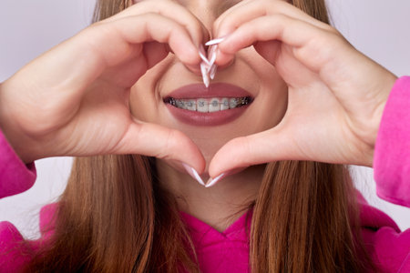 A close-up portrait of a young woman with braces smiling warmly, making a heart shape with her hands. The vibrant pink backdrop adds to the scenes joyful and loving atmosphere.の写真素材