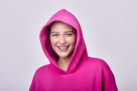 A beautiful young woman with braces on her teeth smiling happily, wearing a trendy pink hoodie. The light pink background complements her joyful expression, creating a vibrant and positive image.の写真素材