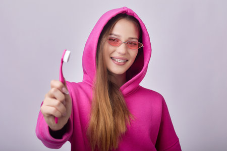 A young woman with braces on her teeth is smiling and holding a toothbrush in her hand. She is wearing a pink hoodie and sunglasses in front of a pink background, showcasing dental care and hygiene.の写真素材