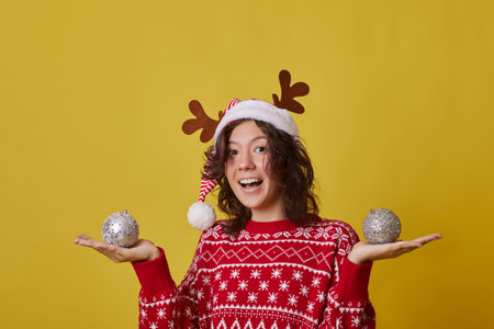 Young woman with curly brown hair smiles in red Santa hat with reindeer antlers, holding silver Christmas balls. She wears cozy red sweater with white snowflake patterns, joyful expression.の写真素材