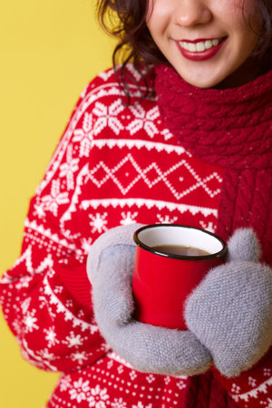 A young woman wearing a red sweater with a white snowflake pattern, a red scarf, and gray gloves is holding a red mug of coffee or tea with both hands. The background is a solid yellow color.の写真素材
