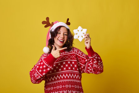 Young woman wearing a red sweater and reindeer antlers headband is smiling while holding a snowflake. She is dressed in festive attire against a bright yellow background.の写真素材