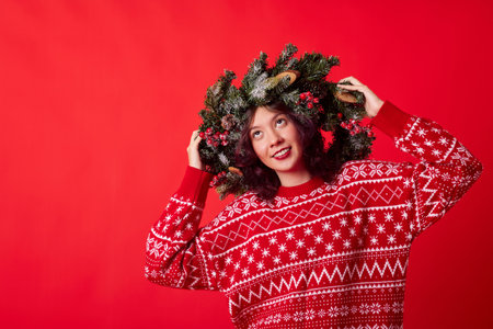 A young woman with long dark hair smiles warmly, wearing a cozy red sweater with white snowflakes and a festive Christmas wreath on her head, spreading holiday cheer.の写真素材
