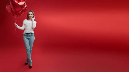 Young woman in casual style wear holds bunch of balloons on red background.の写真素材