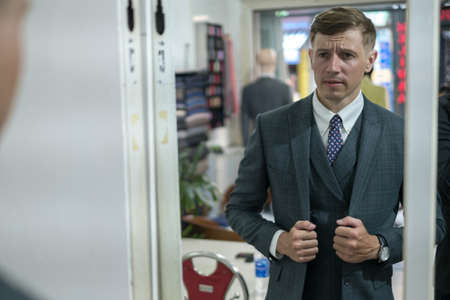 Handsome young businessman trying on new suit in atelier and looking at the mirror. Wondering face. High quality photoの写真素材