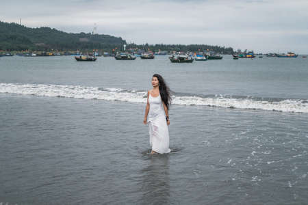 Charming young mongol woman in white dress walking on the beach. Black long curly hair. Looking aside. Romantic photo. Ocean view. High quality photoの写真素材