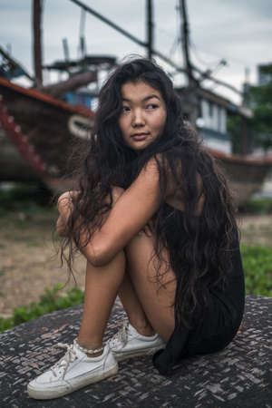 Sitting on the stone near old wooden ships. Blurred background. Black long curly hair. Romantic photo. High quality photoの写真素材